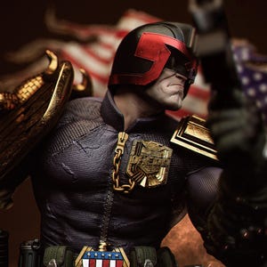 May include: A detailed action figure of Judge Dredd, wearing a black helmet with red accents and a dark blue uniform. The figure has gold shoulder pads and a badge with the American flag. It is holding a gun.