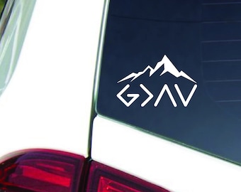 May include: White vinyl decal with the words "God is Greater Than" and a mountain range graphic. The decal is on the back window of a white car.