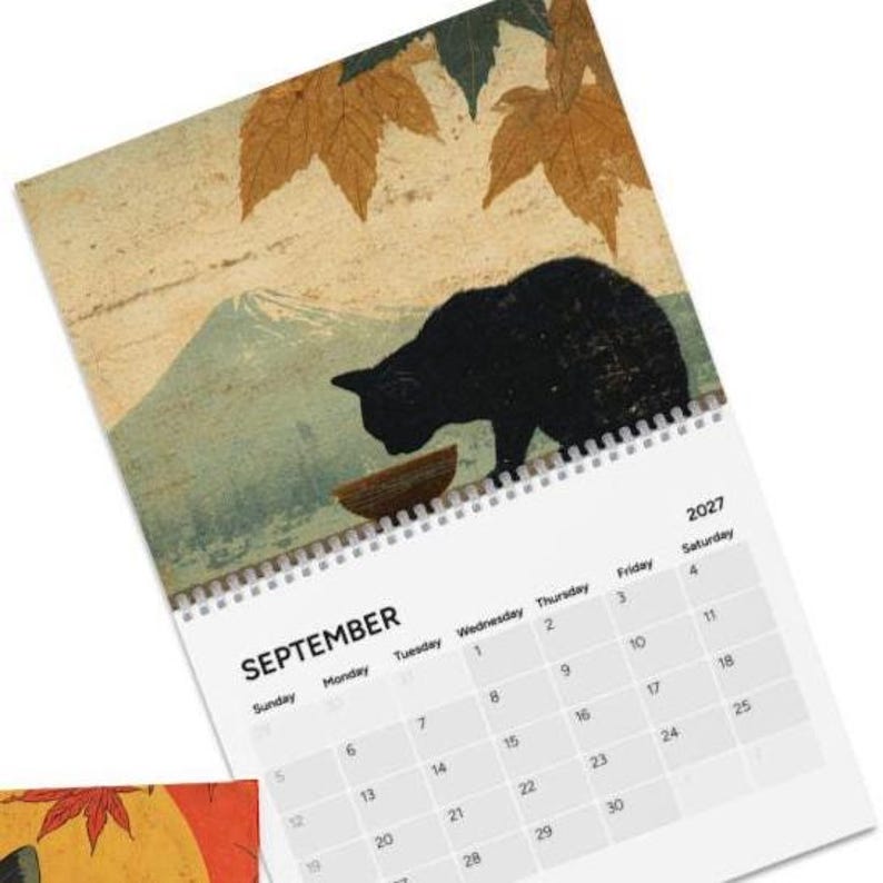 May include: A 2027 wall calendar for September featuring a black cat silhouette drinking from a small bowl. The background displays a mountain landscape with autumn leaves in shades of brown and green. The calendar grid shows dates.