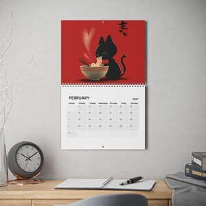 May include: A wall calendar for February 2027 featuring a graphic illustration of a black cat eating noodles from a bowl on a red background. The calendar grid displays the days of the month clearly with black text on a white background.