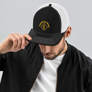 Puede incluir: Una gorra de camionero negra y blanca con un diseño bordado en amarillo que dice "Happiness Ahead" con un símbolo de camino sinuoso.