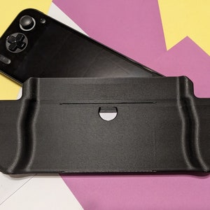May include: A black handheld gaming console with a black protective case. The case has two grips on the sides and a cutout for the console's screen.