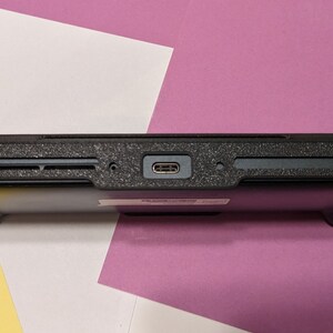 May include: Black handheld gaming console with textured black rubber grips on the sides. The console has a USB-C port on the bottom.