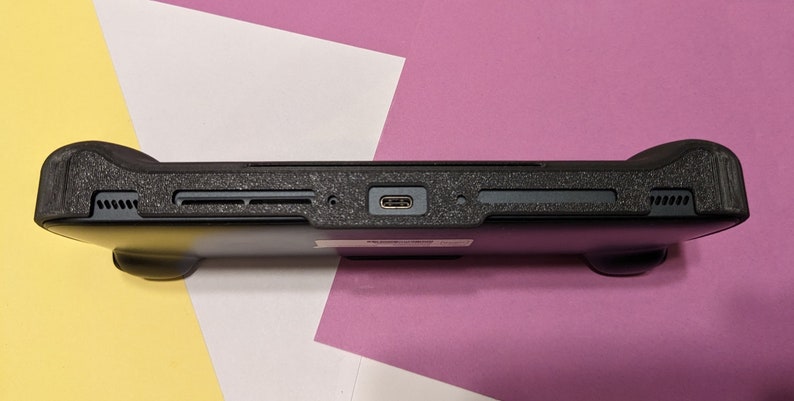 May include: Black handheld gaming console with textured black rubber grips on the sides. The console has a USB-C port on the bottom.