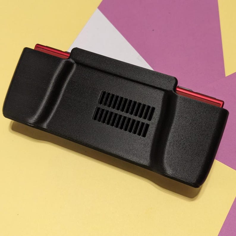 May include: Black plastic case with a red trim and a vent on the back. The case is designed to hold a handheld gaming device.