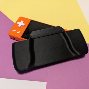 Peut inclure: Un étui noir pour console de jeu portable avec une console de jeu portable orange vif à l'intérieur. La console a un bouton blanc et un écran.