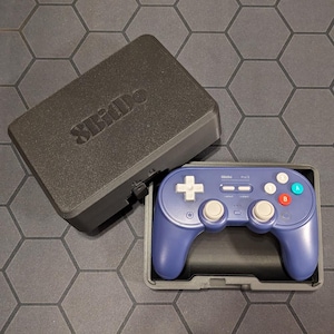 May include: A blue and gray video game controller with a matching case. The controller has white, blue, red, and green buttons. The case is black and has the text "SBido" on the top.