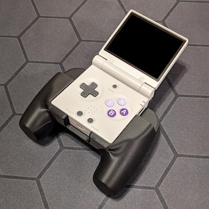 Può includere: Console per videogiochi portatile grigia con schermo nero. La console ha un D-pad nero, pulsanti A e B viola e un'impugnatura nera attaccata in basso. Si trova su una superficie grigia con un motivo esagonale.