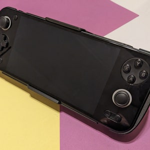 May include: A black handheld gaming device with a screen, buttons, and a joystick. The device is on a pink and yellow surface.