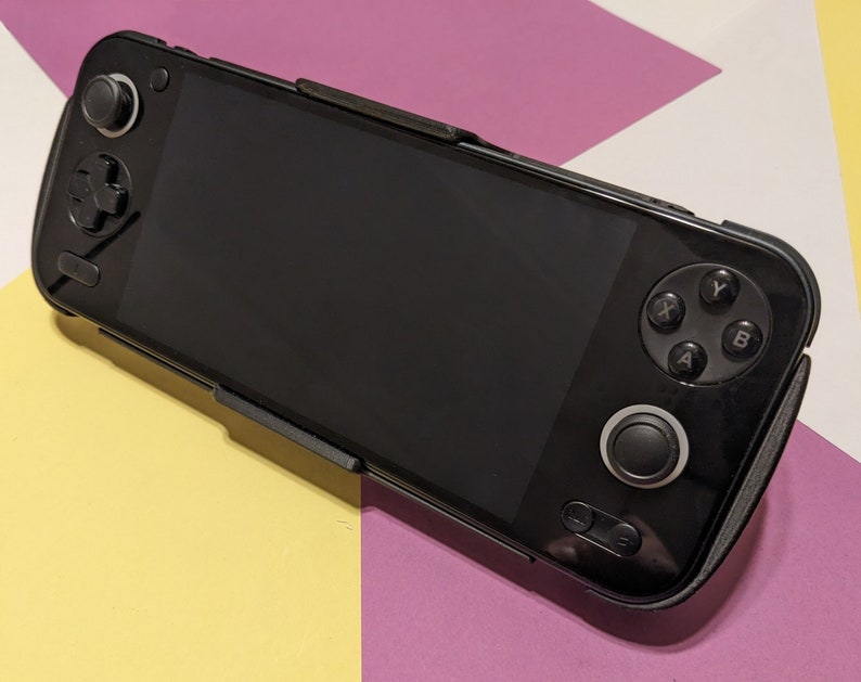 May include: A black handheld gaming device with a screen, buttons, and a joystick. The device is on a pink and yellow surface.