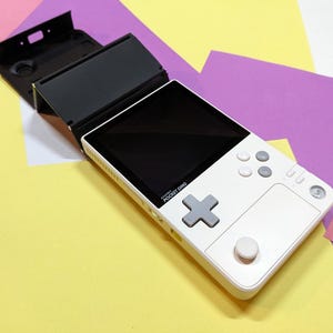 Puede incluir: Una consola de juegos portátil blanca con una pantalla negra y una cubierta negra con bisagras. Cuenta con una cruceta gris, botones grises y un joystick analógico blanco. El texto "AYANEO POCKET DMG" está impreso en el dispositivo.