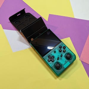 Puede incluir: Consola de juegos portátil verde azulado con botones negros y una pantalla negra, mostrada con su funda protectora negra. El fondo es una mezcla de papel amarillo, morado y rosa. La carcasa es transparente.