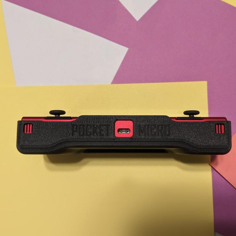 May include: Black handheld gaming console with red accents and the text "POCKET MICRO" printed on the front. The console has two buttons on the top and a USB-C port on the side.