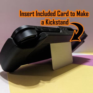May include: A black handheld gaming console with a white kickstand. The kickstand is made from a card that is inserted into the console. The text "Insert Included Card to Make a Kickstand" is visible in the image.