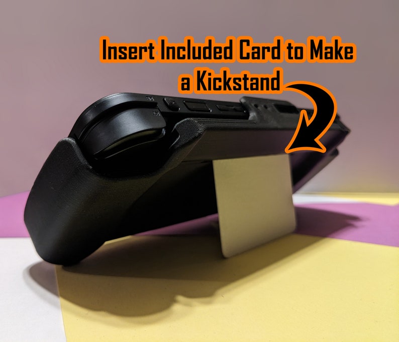 May include: A black handheld gaming console with a white kickstand. The kickstand is made from a card that is inserted into the console. The text "Insert Included Card to Make a Kickstand" is visible in the image.