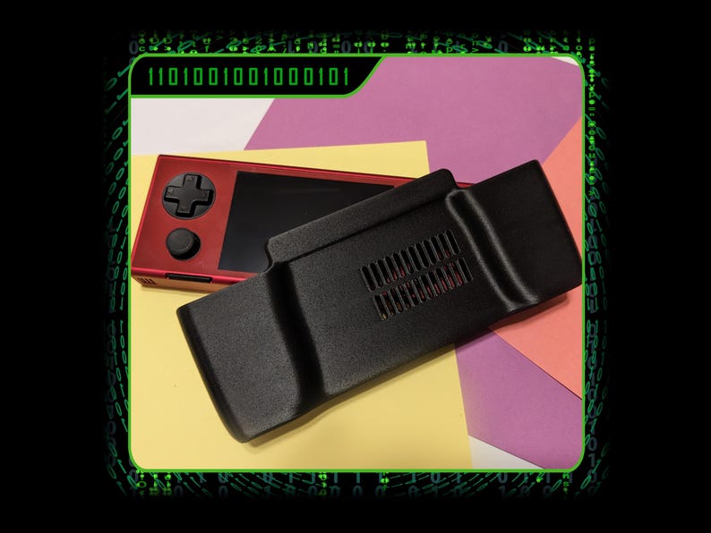 May include: A red and black handheld gaming console with a black case and a red button. The console is on a yellow surface with a pink and orange surface behind it.
