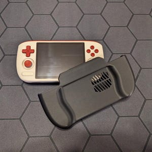 May include: A handheld gaming console with a cream-colored body and red accents on the buttons and directional pad. A separate black grip is positioned nearby. The console features a screen and a sleek, modern design.