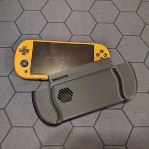 Könnte beinhalten: Eine orangefarbene Handheld-Spielkonsole mit schwarzem Bildschirm und Bedienelementen, neben einer schwarzen Schutzhülle. Auf der Hülle steht das Wort "ALIENWARE".