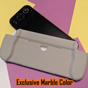 May include: A gray, textured case for a handheld gaming console. The case has a cutout for the console's screen and buttons. The text "Exclusive Marble Color" is printed on the case.