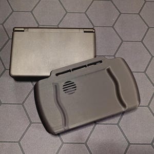 Puede incluir: Dos dispositivos de juegos portátiles negros se muestran sobre una superficie gris con patrón hexagonal. Un dispositivo está cerrado, es rectangular y tiene una bisagra. El otro es un controlador negro con un altavoz y ranuras.
