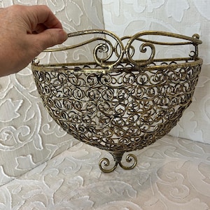 May include: A gold-toned metal wall hanging basket with a decorative scroll design. The basket has a rounded shape and a hanging loop.