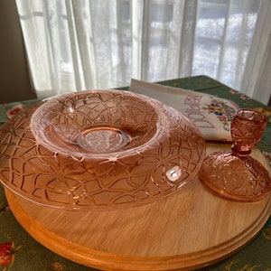 May include: A pink glass pedestal cake stand with a decorative pattern. The stand is made of glass and has a wooden base. The stand is on a green tablecloth with a floral pattern.
