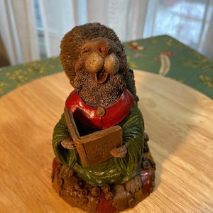 May include: A brown and green squirrel figurine holding a book titled "Carols of the Seasons". The squirrel is wearing a red and green robe and is sitting on a colorful base.