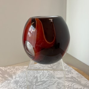 May include: A round glass vase with a swirl pattern in shades of brown, red, and white. The vase is sitting on a clear acrylic stand.