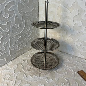 May include: A three-tiered silver-toned serving stand. Each circular tier has a decorative, openwork design. The stand has a central pole with a pointed top. The stand is suitable for displaying desserts or small items.