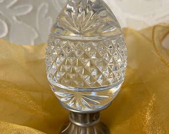 May include: A clear, cut-glass decorative egg-shaped container with a silver-colored base. The egg has a diamond pattern on the lower half and a starburst design on the top. The container sits on a decorative base.