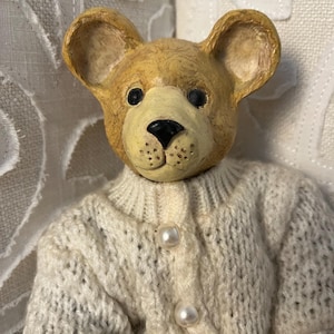 May include: A handcrafted teddy bear figurine with a light brown head and a cream-colored, button-down sweater. The bear has black eyes and a black nose. The sweater has three pearl-like buttons.