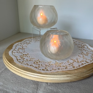 May include: Two frosted glass candle holders with a textured surface, each holding a lit tea light candle. The candle holders are on a wooden tray with a white doily.