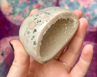 May include: A geode, held in a hand, with a light-colored exterior speckled with green and pink hues. The interior reveals a cavity filled with sparkling crystals. The background is a blurred, colorful pattern.