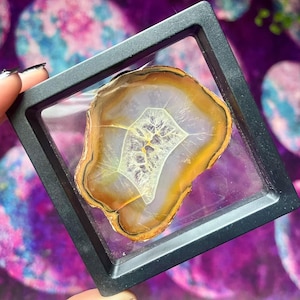 May include: A framed agate slice with a translucent center and bands of white, yellow, and brown. The stone is encased in a black square frame, held against a purple and blue background.