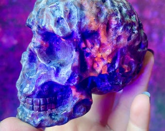 May include: A carved skull sculpture with a textured surface, showcasing shades of purple, blue, and orange. The skull's features are detailed, with visible teeth and eye sockets. The background is a blurred mix of purple and pink.