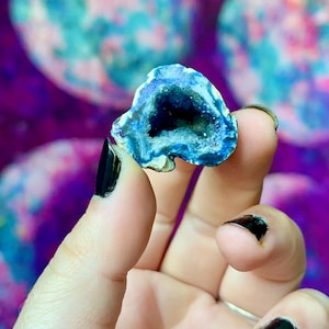 May include: A small, blue geode with a dark interior is held up against a blurred, colourful background. The geode has a crystalline structure and a rough, textured surface. The background features shades of purple, pink, and teal.