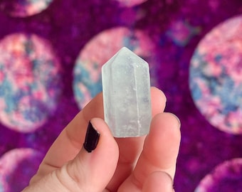 May include: A clear, translucent crystal point with a pointed tip and a rectangular base. The crystal is held in a hand against a purple background with blurred circles.