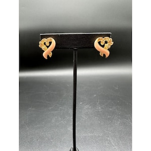 May include: A pair of gold-toned earrings with a ribbon design. The earrings have a light pink enamel finish and are accented with small, clear crystals. The earrings are displayed on a black earring stand against a dark background.