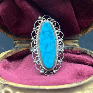 Op de afbeelding: Een zilveren ring met een ovale, heldere turkooizen steen. De ring heeft een gedetailleerd, opengewerkt ontwerp rond de steen. De ring wordt getoond in een vintage sieradendoos met een bordeauxrode fluwelen voering.