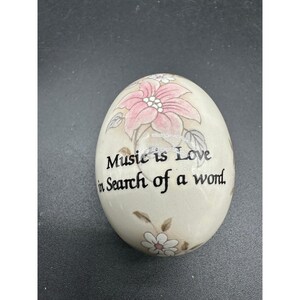 以下が含まれることがあります： クリーム色の卵型の装飾品。ピンクと白の花柄のデザイン。テキスト「Music is Love in Search of a word」は黒で印刷されています。ユニークで芸術的な作品。