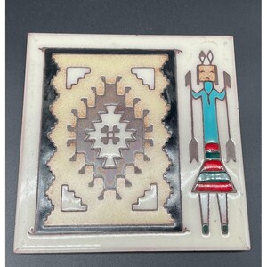 May include: Square ceramic tile with a Southwestern design. The tile features a central geometric pattern in beige, brown, and white, framed by a black border. A stylized figure with turquoise, red, and brown accents is on the right.