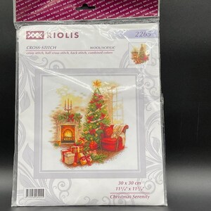 May include: A Riolis cross-stitch kit depicting a Christmas scene. The design includes a decorated Christmas tree, a fireplace, a red armchair, and wrapped gifts. The kit measures 30 x 30 cm (11 3/4" x 11 3/4"). The text on the package reads "Christmas Serenity".