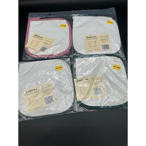 Puede incluir: Cuatro agarraderas blancas con ribetes de colores en rosa, blanco y verde, cada una empaquetada en plástico transparente. Cada agarradera tiene una etiqueta que dice "Janlynn Personal Wares Potholder".