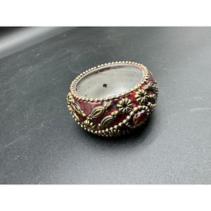 Puede incluir: Un brazalete decorativo con acabado de esmalte rojo y detalles metálicos dorados. El brazalete presenta diseños florales y de hojas, con una piedra central de color ámbar. El brazalete tiene aproximadamente 6,35 cm de diámetro.