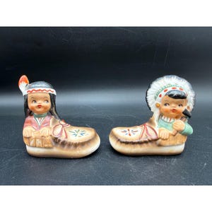 Può includere: Due statuette vintage in ceramica raffiguranti bambini nativi americani. Ogni figura è seduta in una base a forma di mocassino. Uno indossa un copricapo di piume e l'altro un copricapo di piume bianche. Le statuette sono dipinte con colori vivaci.