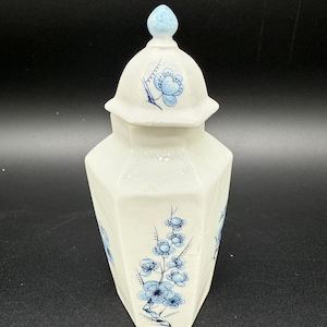 May include: A white ceramic jar with a blue floral design. The jar has a lid with a small knob on top. The jar is decorated with blue flowers and leaves.