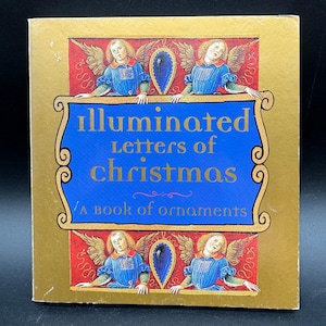 Puede incluir: Un libro cuadrado con una cubierta dorada y un panel azul con el texto "illuminated Letters of Christmas". La portada del libro tiene ilustraciones de ángeles y adornos decorativos. También es visible el texto "A book of ornaments".