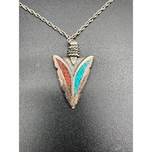 Könnte beinhalten: Silberfarbene Pfeilspitzen-Anhänger-Halskette mit roten und türkisfarbenen Einlagen. Der Anhänger hat ein strukturiertes Design und hängt an einer filigranen Kette. Ein einzigartiges Schmuckstück.