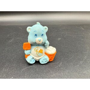 May include: A light blue Care Bear figurine with a baby-themed design. The bear is holding an orange shovel and a small orange bucket filled with white material. The bear has a white diaper with a baby carriage design.