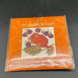 May include: An orange cross-stitch card kit featuring a red dormouse with purple berries. The kit is titled "Dormouse" and "Countryside Miniatures". The packaging is orange with a clear plastic wrap.